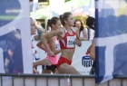 Na pierwszym planie jest kobieta, pokazana bokiem w trakcie biegu, ubrana w czerwony strój. Po obu stronach zdjęcia są dwie niebieskie flagi 