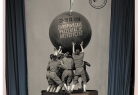 Plakat prezetnuje mężczyzn stojących na fortepianie, trzymających dużą kulę, na której znajduje się tancerka baletowa, z jedną podniesoną nogą i trzymająca w dłoni różę. W tle napis pępek świata, plakat stylizowany na lata 30. XX wieku