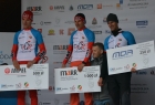 czterech mężczyzn stoi na podium na tle planszy z logami sponsorów. Trzech z nich trzyma duże czeki z kwotą za wywalczone miejsce