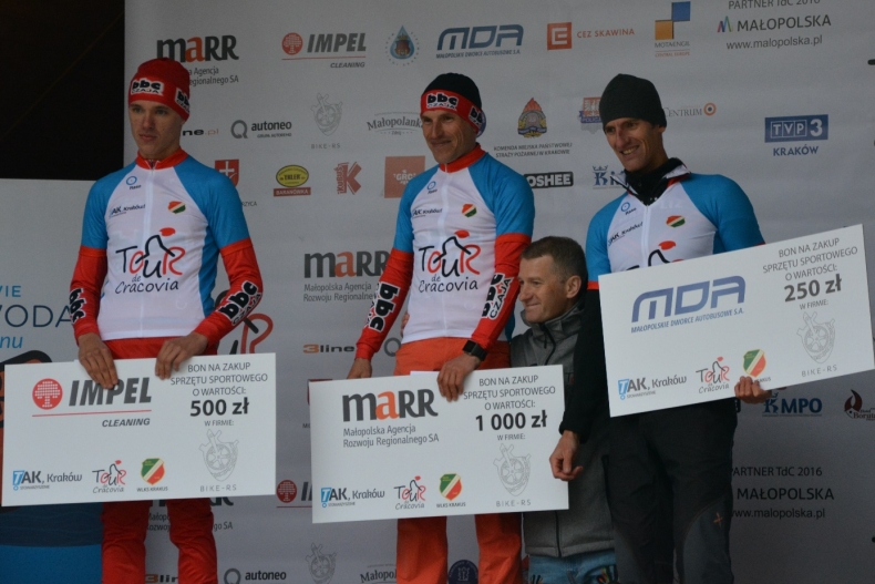 czterech mężczyzn stoi na podium na tle planszy z logami sponsorów. Trzech z nich trzyma duże czeki z kwotą za wywalczone miejsce