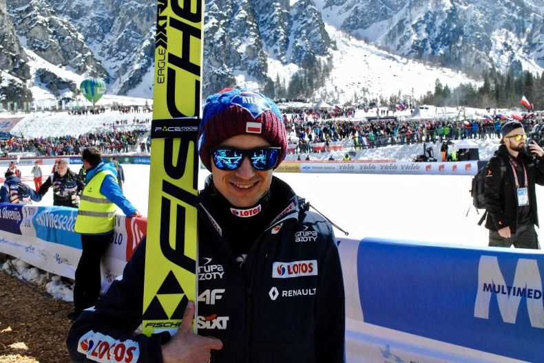 sportowiec z nartami wzdłuż jego ciała, w czapce i okularach przeciwsłonecznych stoi na tle ośnieżonych gór