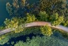 Zdjęcie przedstawia ujęcie z góry (z drona) trasy rowerowej biegnącej przez stawy i las