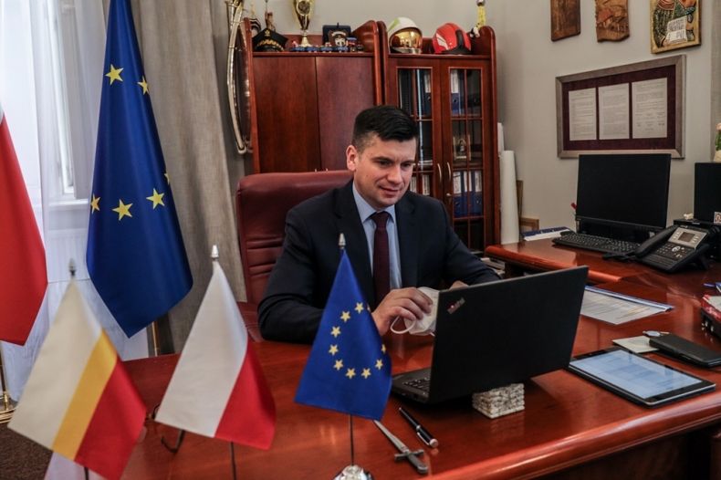 mężczyzna w czarnym garniturze siedzi przed ekranem komputera, ustawionym na biurku, obok niego stoja trzy flagi, Polski, Województwa Małopolskiego i Unii Europejskiej
