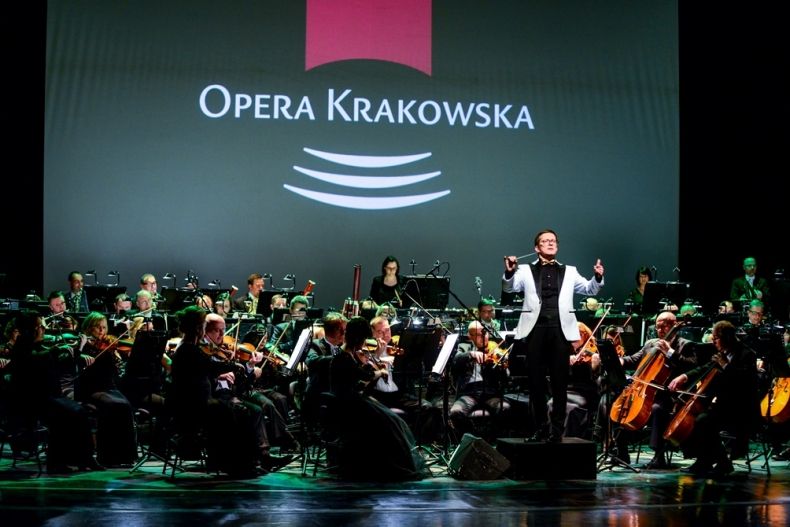 Orkiestra Opery Krakowskiej pod dyrekcją Tomasza Tokarczyka w trakcie występu. Artyści, siedzący na scenie w czterech półkolistych rzędach, grają na instrumentach smyczkowych i dętych. Na pierwszym planie, frontem do publiczności, dyrygent z uniesioną bat