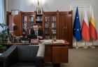 Widok z oddali: wicemarszałek Tomasz Urynowicz siedzi przy biurku w swoim gabinecie, w tle biblioteczka oraz trzy flagi