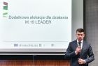 Wicemarszałek Łukasz Smółka trzyma mikrofon w ręce i przemawia. Z tyłu wyświetlony slajd z napisem Dodatkowa alokacja na działania M.19 LEADER.