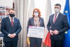 Wręczenie promesy na dofinansowanie dla gminy Świątniki Górne, dwaj mężczyźni i kobieta stoją na tle flag