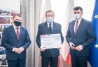 Wręczenie promesy na dofinansowanie dla gminy Trzyciąż, trzej mężczyźni stoją na tle flag