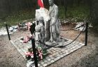 Pomnik, postać człowieka z kamienia, obok złożone wieńce, w tle biało czerwona flaga.