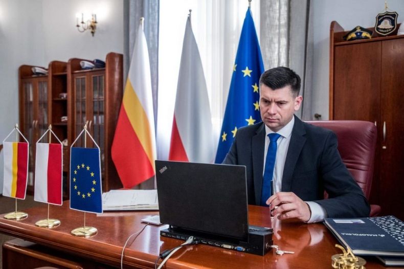 Wicemarszałek Łukasz Smółka siedzi w gabinecie przed komputerem. Obok widoczne flagi Małopolski, Polski i Unii Europejskiej. Z tyłu meble.