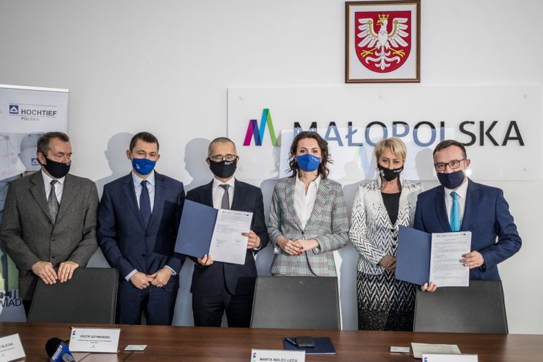 Grupa uczestników konferencji prasowej stojąca na tle logo Małopolski z podpisanymi umowami