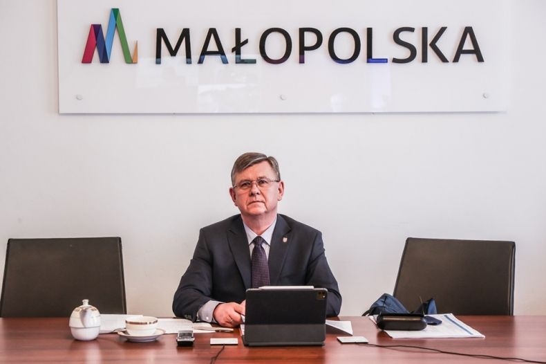 Marszałek Witold Kozłowski siedzi za biurkiem. Na biurku tablet, filiżanka i dokumenty. W tle napis Małopolska.