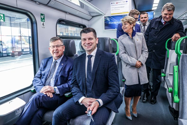 Marszałek Witold Kozłowski i wicemarszałek Łukasz Smółka siedzą w nowym pociągu w kabinie pasażerskiej. Z tyłu widoczni podróżni.