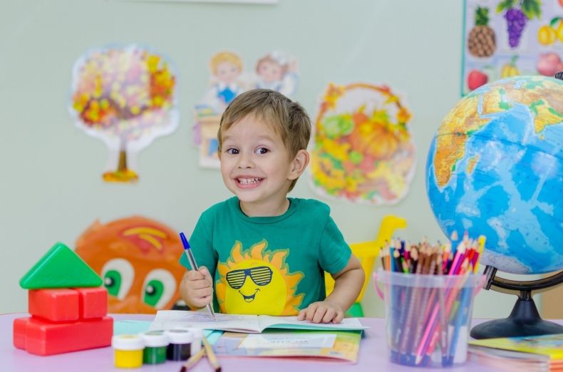 Uśmiechnięte dziecko siedzi przy biurku, na którym stoi globus, a także znajdują się kredki, kartki papieru oraz inne kolorowe przybory szkolne.