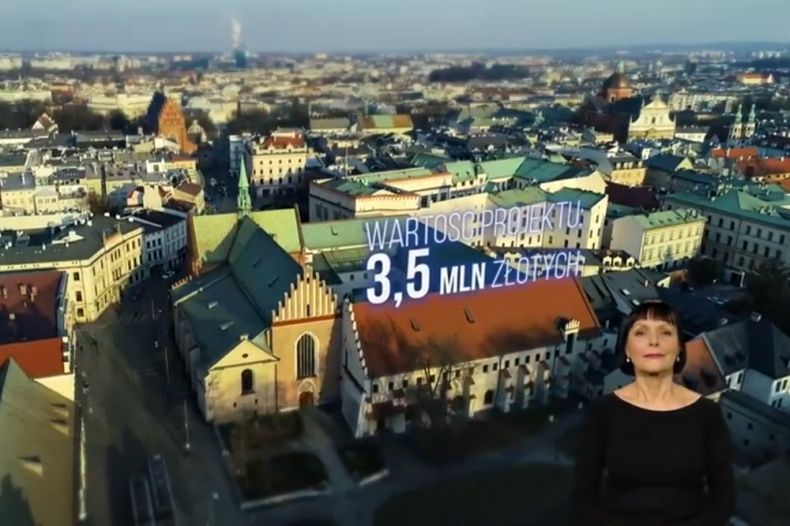 Ujęcie z filmu - widok z lotu ptaka na kościół i miasto, na środku zdjęcia napis "wartość inwestycji: 3,5 mln zł". W prawym dolnym rogu postać tłumaczki na język migowy
