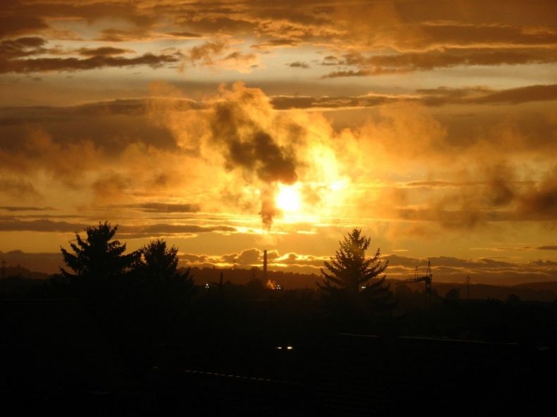 Dym z kominów na tle zachodzącego słońca nad zabudowaniami i koronami drzew