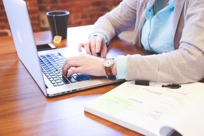 Osoba siedząca przy otwartym laptopie, zbliżenie na dłonie piszące na klawiaturze, na biurku leży notes, w tle kubek z herbatą