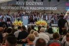Widok na scenę , na której stoją przemawiający ludzie. Nad sceną widać napis 24 dożynki województwa małopolskiego. W tle widać tłumy ludzi stojących pod sceną. 
