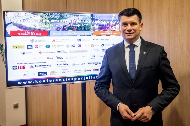 Wicemarszałek Łukasz Smółka stoi na tle telewizora z wyświetloną planszą IX Krakowskie Dni Bezpieczeństwa Ruchu Drogowego.