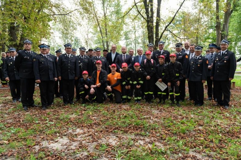 Wspólne pamiątkowe zdjęcie młodych strażaków i zaproszonych gości na tle zieleni w parku