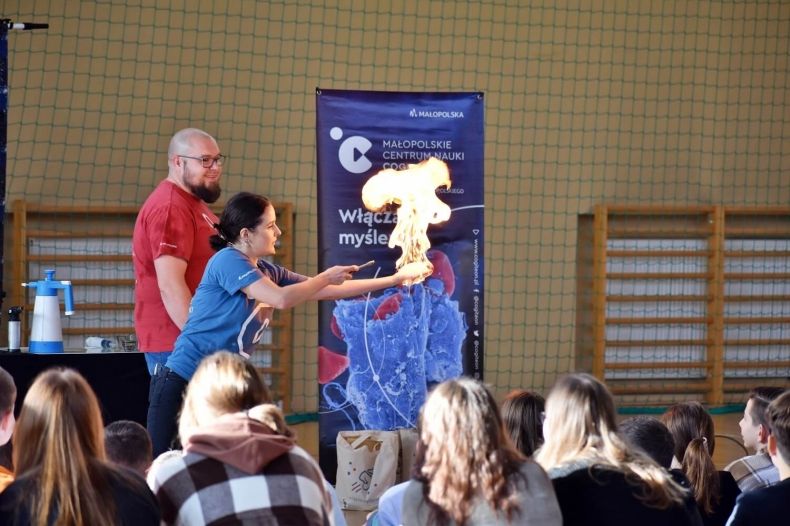 Pokaz naukowy w sali gimnastycznej szkoły. Widać publiczność oraz edukatorkę, która trzyma przed sobą wyciągniętą dłoń a na niej płomień spalanej substancji.