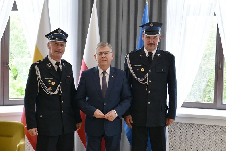 Marszałek Witold Kozłowski i dwóch strażaków w strojach galowych. Mężczyźni stoją na tle trzech flag Małopolskiej, Polskiej i Unijnej. 