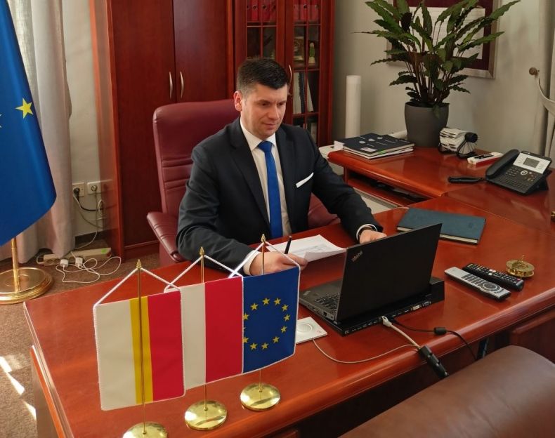 Wicemarszałek Łukasz Smółka siedzi przy stole w gabinecie i patrzy w monitor komputera. Na pierwszym planie widoczne flagi Małopolski, Polski i Unii Europejskiej.