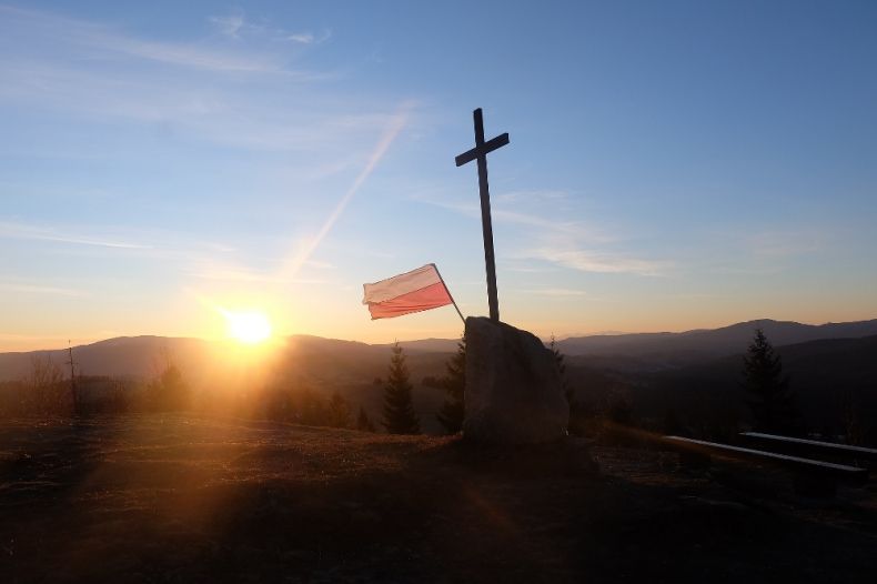Flaga RP i krzyż na tle nieba i wschodzącego słońca
