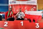 Trzech zawodników na wózkach na podium. Jeden z nich trzyma flagę Polski. Za nimi baner reklamowy IWAS World Games 2022.