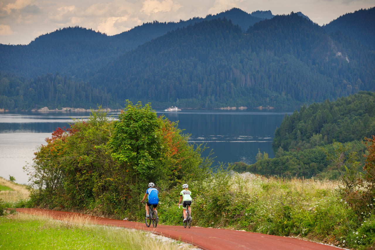 Na zdjeciu widać ścieżkę rowerową jedzie po niej dwóch rowerzystów w tle jezioro i góry