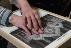 Zdjęcie przedstawia dłonie starszej osoby dotykające eksponat muzealny przystosowany dla osób z dysfunkcją wzroku