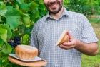 Mężczyzna w jasnej koszuli, na tle winorośli prezentuje miskę chlebową z żurkiem, ułożoną na białym talerzu, który stoi na drewnianej okrągłej desce.