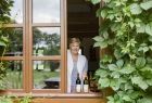 Kobieta w oknie obrośniętym zielonym pnączem prezentuje produkty winnicy.