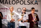trzech mężczyzn, jeden z nich w okularach przeciwsłonecznych, jeden w marynarce, pozstali w koszulach trzymaja w rękach nagrody tuż przed dekoracją 