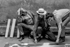 Czarno-biała fotografia przedstawiająca trzech mężczyzn: od lewej: Jacek Stokłosa, Wacław Janicki, Lesław Janicki. Wszyscy kucają. pierwszy od lewej patrzy w górę. Pozostali dwa pochylają się nad grafiką rozłożoną na ziemi
