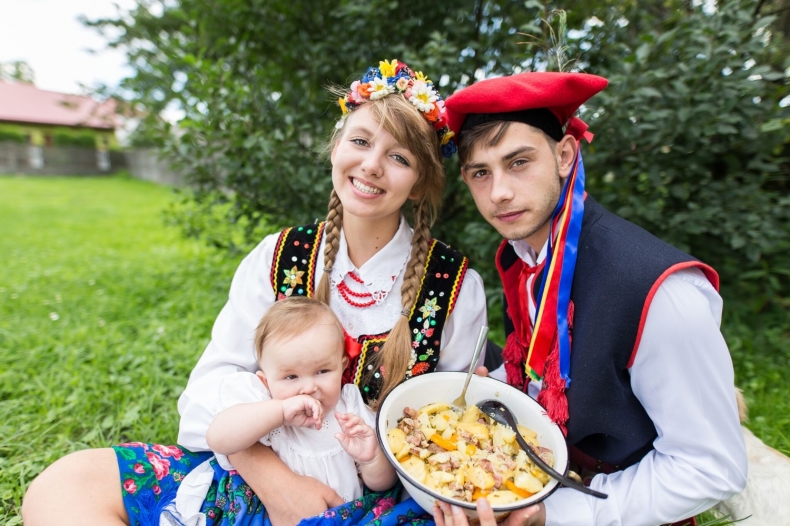Kobieta i mężczyzna w strojach ludowych z dzieckiem. Mężczyzna trzyma emaliowaną miskę z ziemniakami