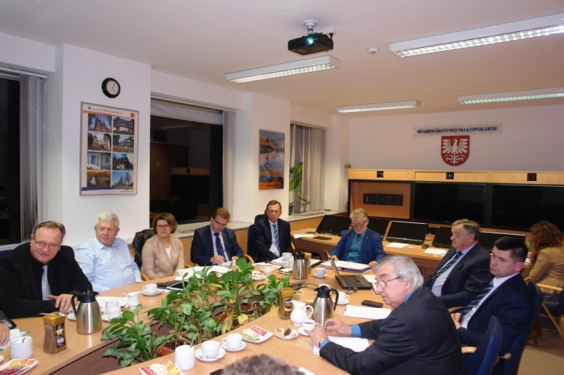 zdjęcie przedstawia osoby siedzące przy owalnym stole, w tle na ścianie herb województwa z podpisem województwo małopolskie