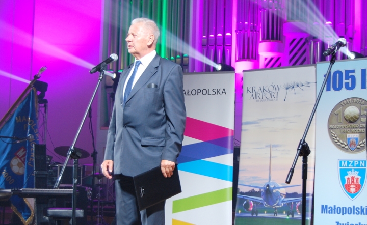 mężczyzna w garniturze na tle rollapów promocyjnych m.in. Małopolski przemawia do mikrofonu na estradzie