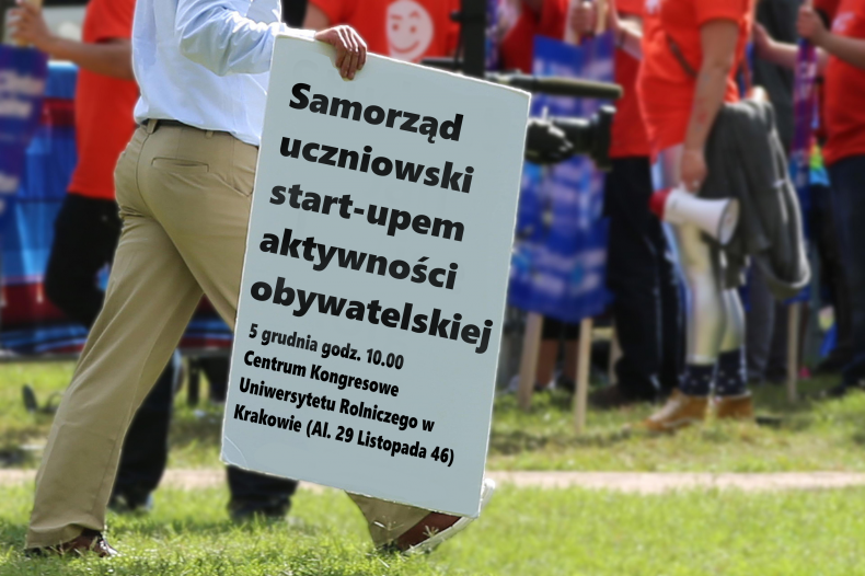 Zdjęcie. mężczyzna idący po zielonej trawie. W ręce trzyma białą tablicę, na której znajduje się napis Samorząd uczniowski start-upem aktywności obywatelskiej, 5 grudnia 2016 r. w Centrum Kongresowe Uniwersytetu Rolniczego (Al. 29 Listopada 46, Kraków