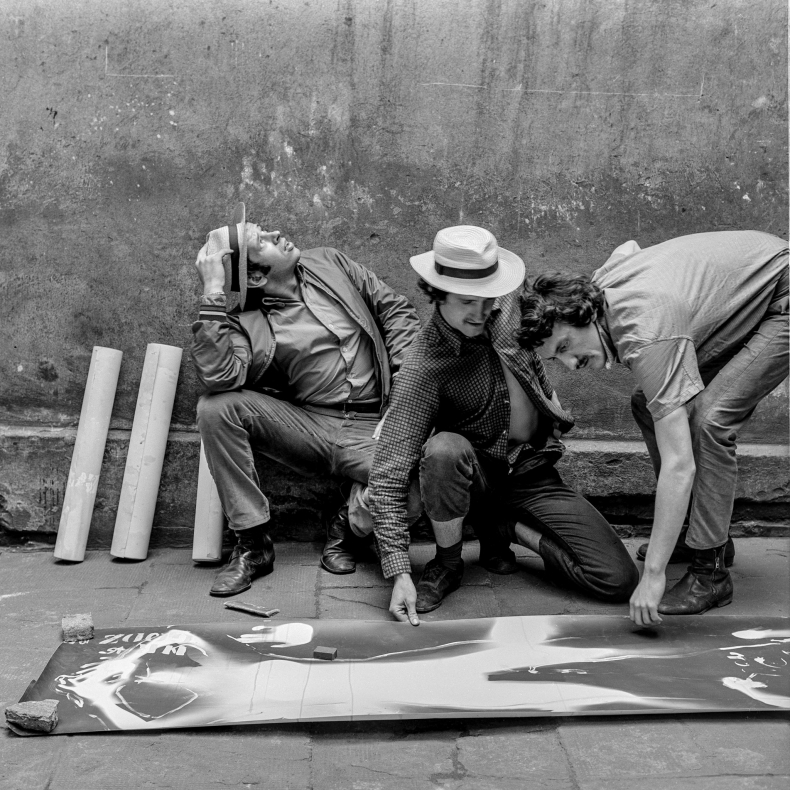 Czarno-biała fotografia przedstawiająca trzech mężczyzn: od lewej: Jacek Stokłosa, Wacław Janicki, Lesław Janicki. Wszyscy kucają. pierwszy od lewej patrzy w górę. Pozostali dwa pochylają się nad grafiką rozłożoną na ziemi