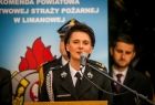 Przewodnicząca Urszula Nowogórska stoi przy mównicy ubrana w mundur strażacki. W tle baner z Państwowej Straży Pożarnej.