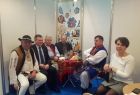 Grupa osób siedzi przy stoliku. Po lewej i po prawej stonie stołu mężczyźni ubrani w stoje ludowe.