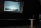 Janusz Sepioł stojący przy mównicy. W tle na ekranie wyświetlany jest slajd.