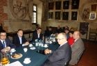 Grupaosób siedząca przy owalnym stole. Na ścianach obrazy i freski.