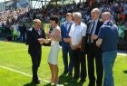 Przewodnicząca Urszula Nowogórska stojąca na murawie w grupie osób wręcza puchar mężczyźnie w garniturze.