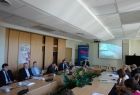 Grupa osób siedzących przy stole. W tle na ścianie wyświetlana jest prezentacja.