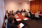 Grupa osób siedzących przy stole w kształcie litery "u". Na ścianie w tel godła Polski i Małopolski.