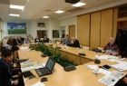 Grupa osób siedzących przy prostokątnym stole. W tle na ścianie obraz przedstawiający pejzarz z drzewem i chmurami.