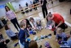 Na zdjęciu w okręgu grupa dzieci i dorosłych, siedzą lub klęczą w sali. tworzą coś z szalika i rękawiczek