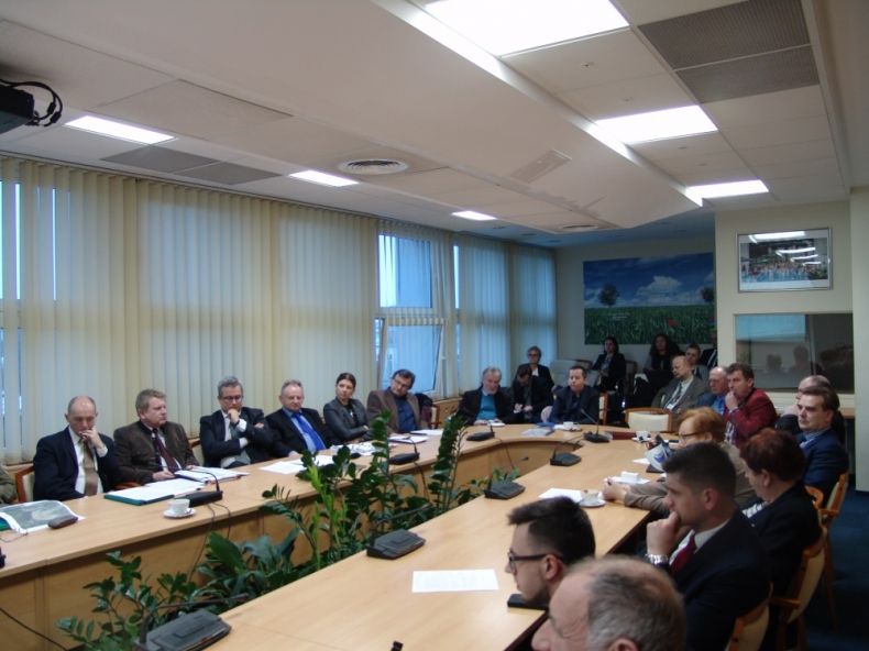 Grupa osób siedząca przy owalnym stole, w tle rząd okien.
